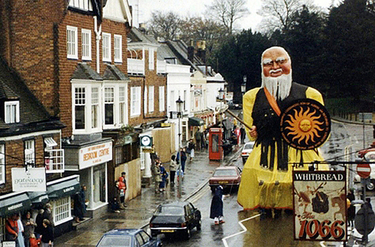 The model of Gog Magog giant is paraded through the town of Battle. He is depicted wearing a black waistcoat over a yellow tunic and carries a round black shield displaying the sun and a crude sword.