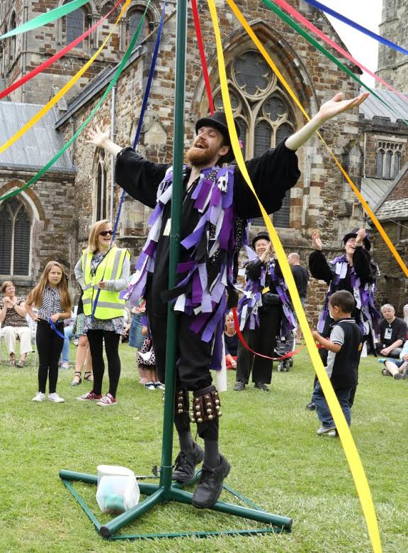 A member of Anonymous Morris is standing at the centre of a maypole dance to stabilise the central pole. We can clearly see his kit as is described by the rest of this article - black hat, shirt, trousers and shoes plus a rag jacket with black, white and purple tatters. Around him we can see children and parents taking part in a maypole dance.