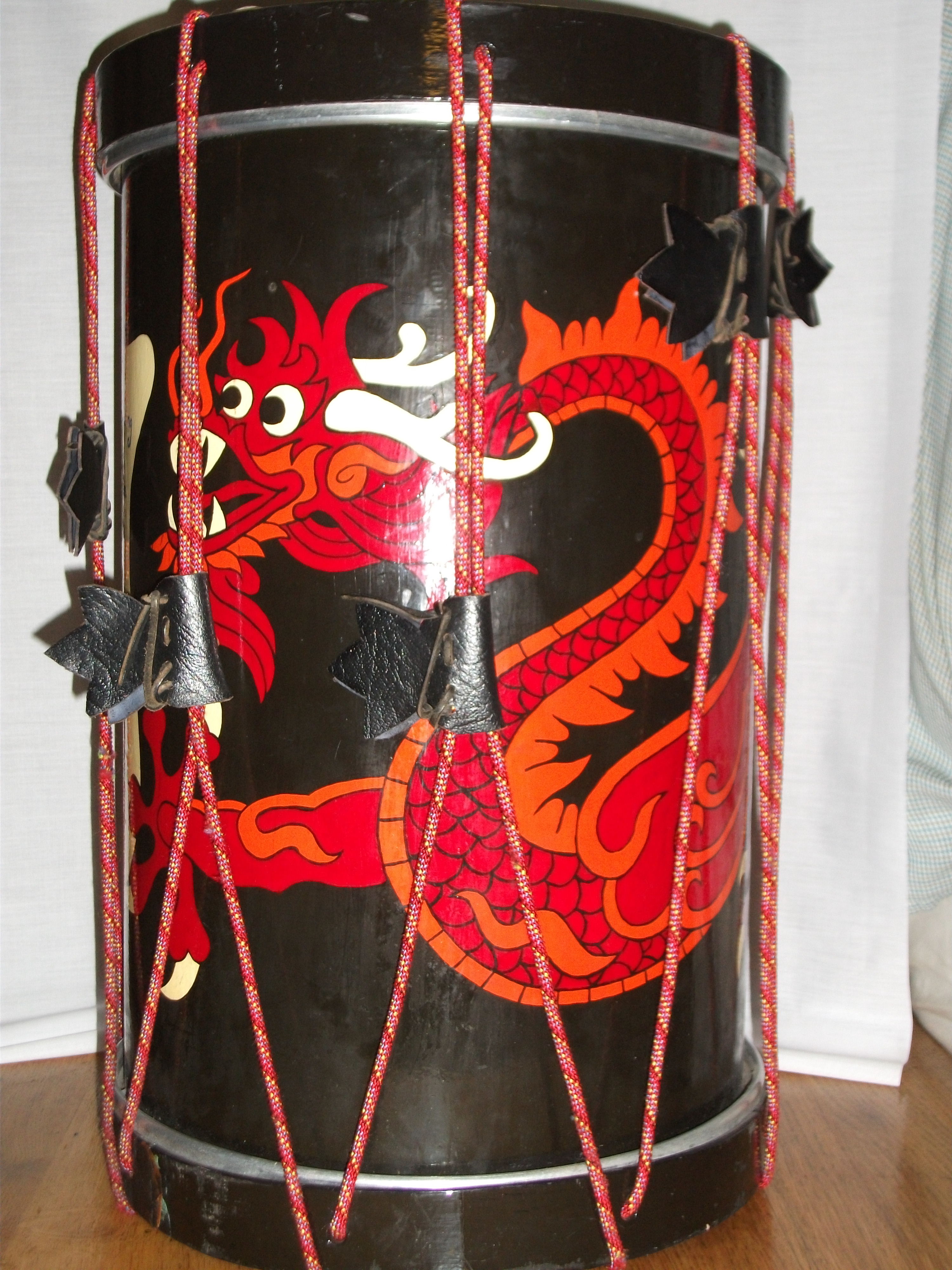Tall drum, painted black with an oriental dragon painted in red circling around the body of the drum. The top and bottom drum skins are kept in tension by red cord running the full height of the drum in a zig-zag pattern.