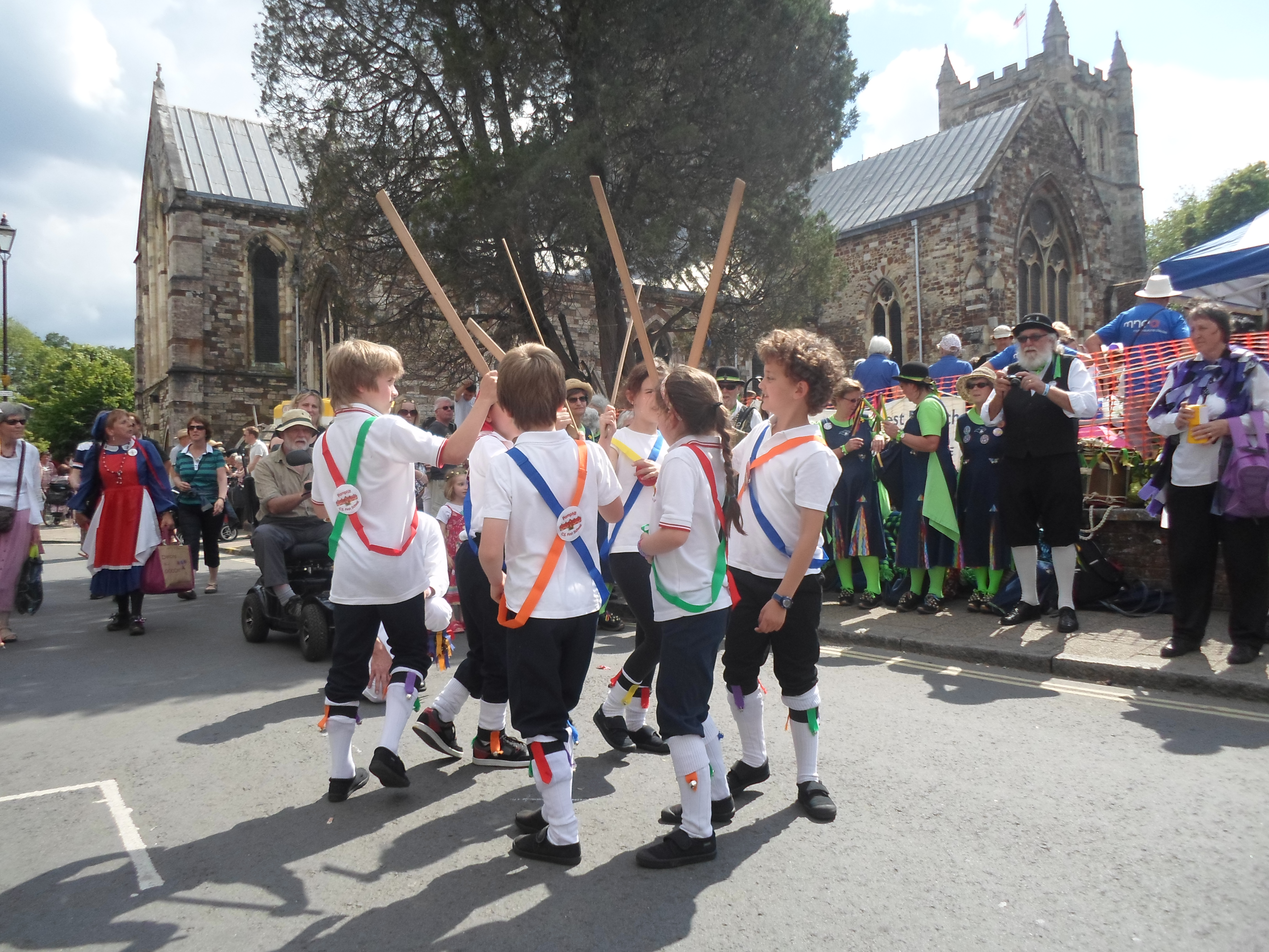 Six children wearing white tops, black trousers and long white socks with ribbons and bells are dancing a longsword dance in a circle, their sticks raised in the air ready to strike in the centre.