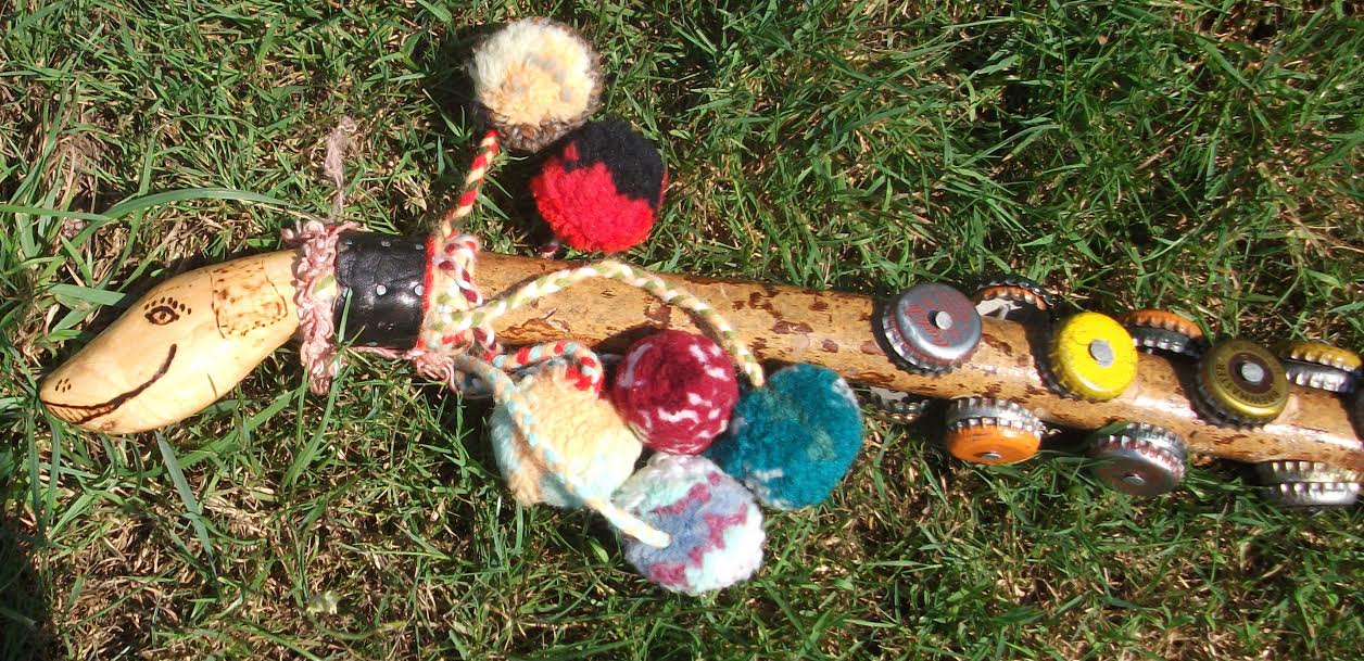 A Mendoza, or Monkey Stick, with a shaped and decorated stylised reptilian head at the top, and several rows of bottle caps nailed down its length. Near the top of the stick, attached to the 'collar' of the head, are a selection of pom-poms on short lengths of yarn.